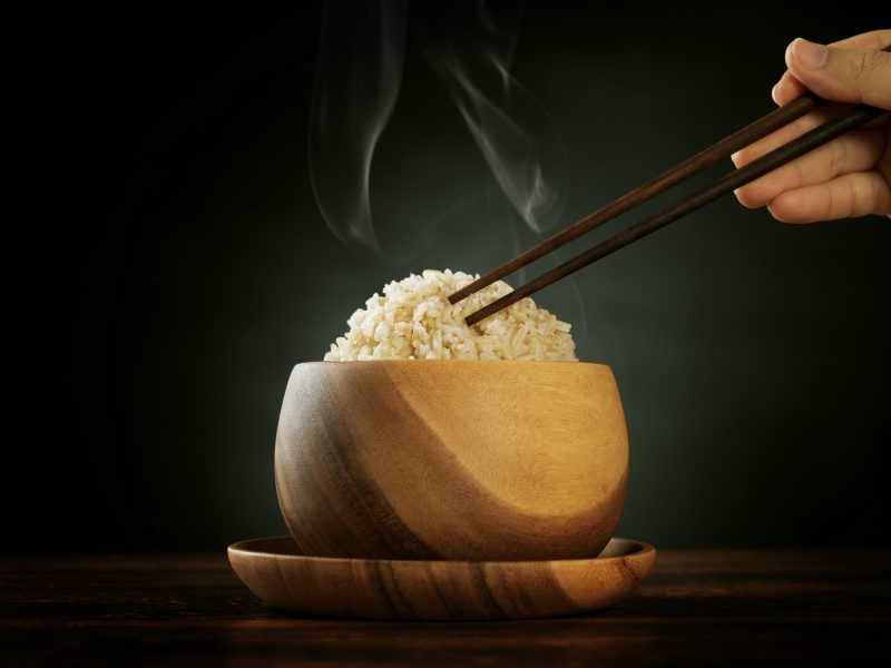 Cooked organic basmati brown rice in wooden bowl with human hand chopsticks and hot steam smoke on dining table. Low light setting.